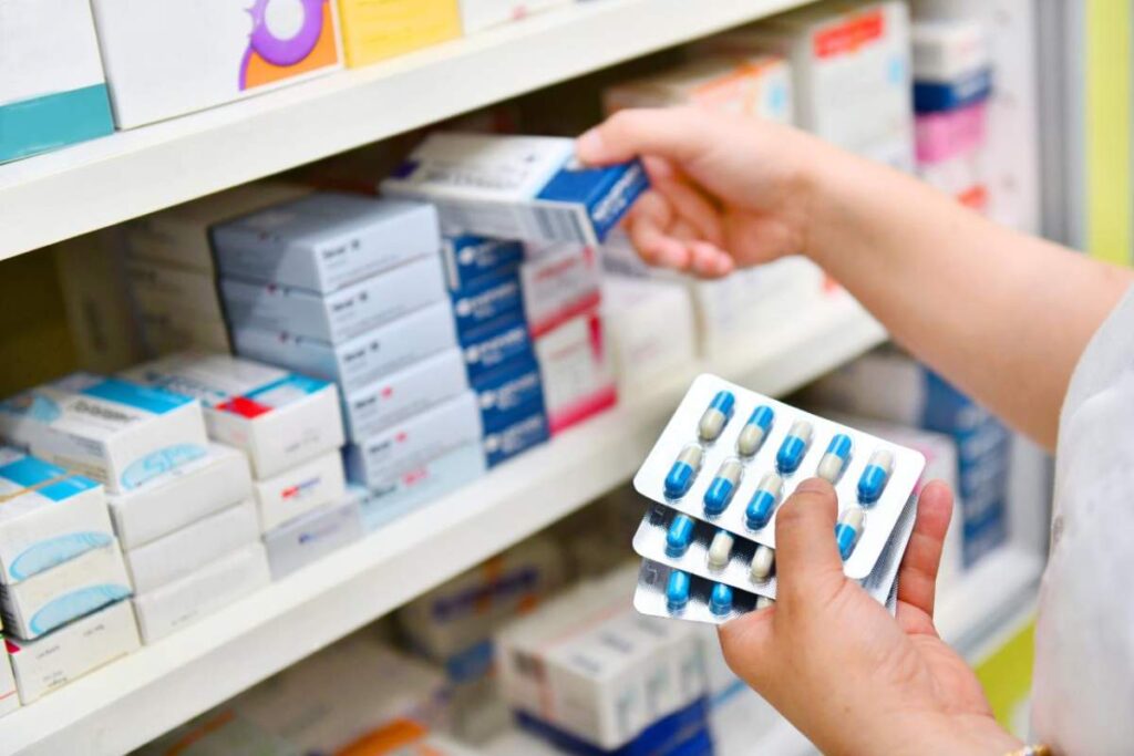 A pharmacist hands a prescription bag to a customer across a pharmacy counter, discussing medication instructions.