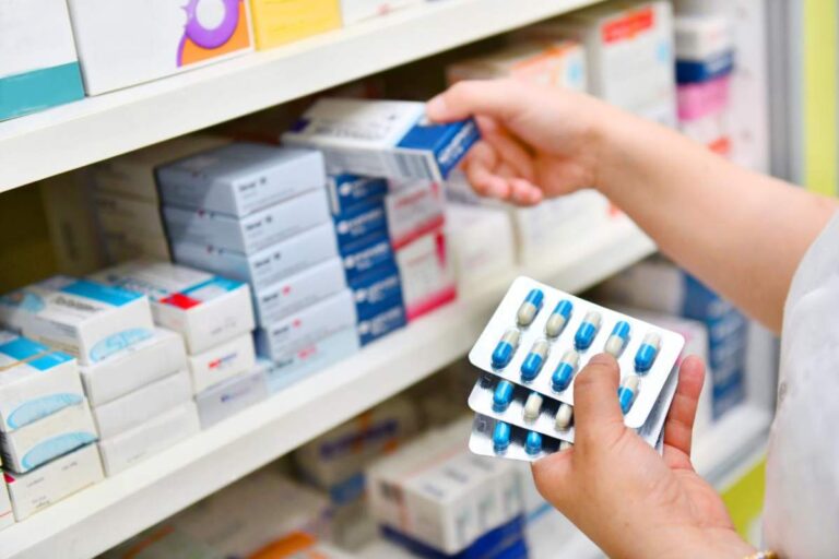 A pharmacist hands a prescription bag to a customer across a pharmacy counter, discussing medication instructions.