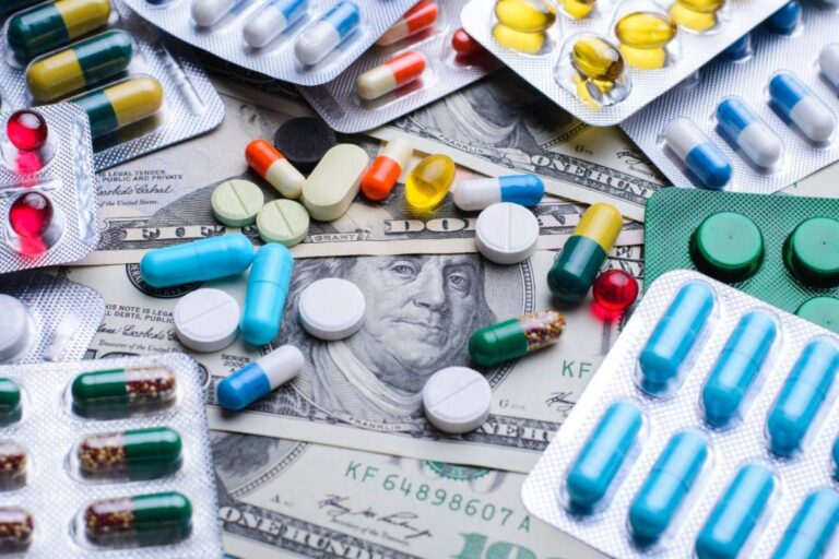 A person checks a medication schedule with a pill organizer and a phone alarm on a kitchen counter.